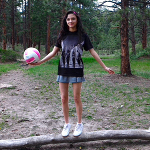 a young woman holds a volleyball and stands on a log. she wears a black t-shirt  with a vintage photo from the 1940s of four women walking carrying skis